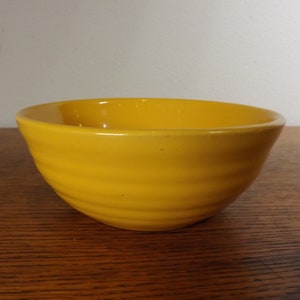 May include: A yellow ceramic bowl with a wavy, ribbed design. The bowl is empty and sitting on a wooden surface.