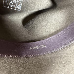 May include: Close-up of a dark olive green fedora hat. The interior features a purple band with the number "A196-732" printed in silver. A tag with text is visible.
