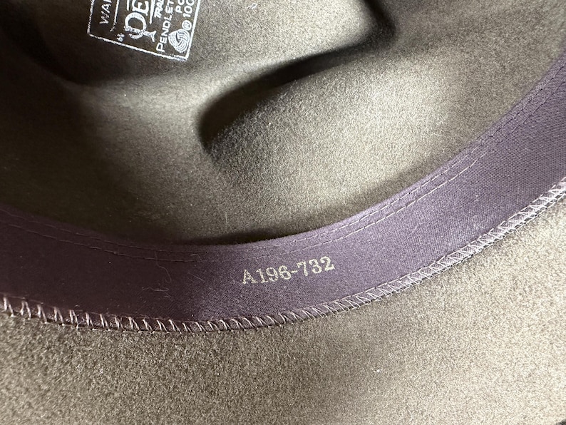 May include: Close-up of a dark olive green fedora hat. The interior features a purple band with the number "A196-732" printed in silver. A tag with text is visible.