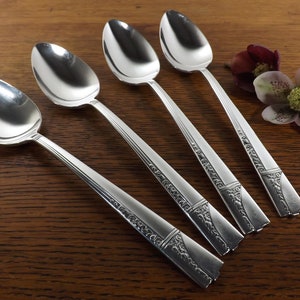 May include: Five silver spoons with ornate, detailed handles are arranged diagonally on a wooden surface. The spoons have a reflective, polished finish. A decorative flower with burgundy and green petals is in the background.