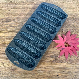 May include: A cast iron cornbread pan with seven corn cob molds. The pan is dark gray and sits on a wooden surface. Red maple leaves are to the right of the pan. The pan has handles for easy use.
