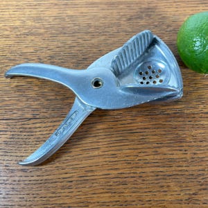 May include: A silver-colored citrus juicer with a hinged design and a perforated bowl. The juicer is next to a green lime. The juicer is made of metal and has a handle for squeezing citrus fruits.