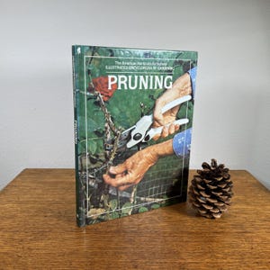 May include: A hardcover book titled "PRUNING" from The American Horticultural Society. The cover features a close-up of hands using pruning shears on a rose bush. A pine cone sits to the right of the book on a wooden surface.