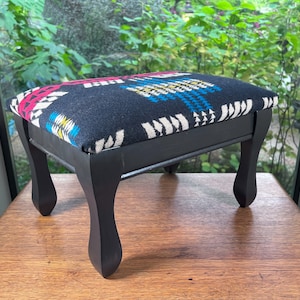 May include: A black footstool with a colorful geometric patterned fabric top. The footstool has four legs and is painted black.
