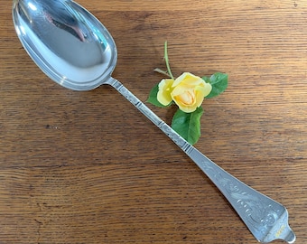 May include: A silver serving spoon with an oval bowl and ornate handle. The handle features engraved floral designs and geometric patterns. A yellow rose with green leaves rests near the spoon. The spoon is on a wooden surface.