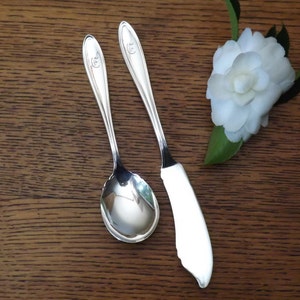 Può includere: Due utensili d'argento, un coltello da burro e un cucchiaio, su una superficie di legno. Il coltello ha una lama curva e il cucchiaio ha una ciotola arrotondata. Entrambi gli utensili hanno un design decorativo sul manico.