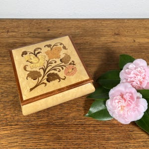 May include: A square wooden jewelry box with a floral inlay design in shades of yellow, brown, and pink. The box is light-colored with a dark brown border. Two pink flowers with green leaves are next to the box.