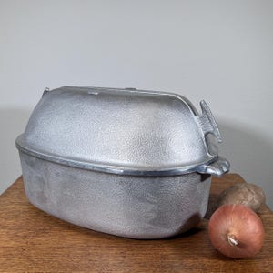 May include: A vintage aluminum roasting pan with a textured surface and a lid. The pan has handles on the sides and a handle on the lid. An onion and two stones are next to the pan on a wooden surface.