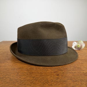May include: A brown fedora hat with a black grosgrain ribbon band. The hat has a classic, structured shape with a slightly curved brim. The hat is sitting on a wooden surface.