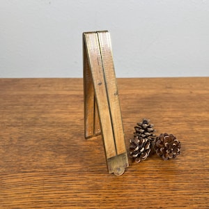 May include: A vintage wooden folding ruler with brass accents, partially open, resting on a wooden surface. Two small pinecones are next to the ruler. The ruler appears to be well-worn, with visible measurement markings.
