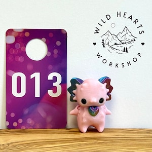 May include: A pink figurine with a heart-shaped pendant and colorful accents stands next to a purple tag with the number 013. The Wild Hearts Workshop logo is visible in the background.