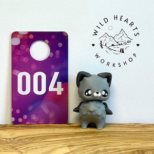 Könnte beinhalten: Eine graue, cartoonartige Katzenfigur mit großen Augen und kleinen Flügeln, neben einem lila-weißen Anhänger mit der Nummer 004. Das Logo von Wild Hearts Workshop ist im Hintergrund zu sehen.