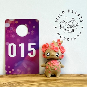 May include: A tan figurine with pink floral accents and black eyes stands next to a purple tag with the number 015. The figurine has a whimsical design, and the tag has a bokeh effect. The Wild Hearts Workshop logo is also visible.