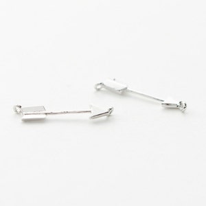 Könnte beinhalten: Zwei silberne Pfeil-Anhänger mit schlichtem Design. Die Pfeile zeigen in entgegengesetzte Richtungen.