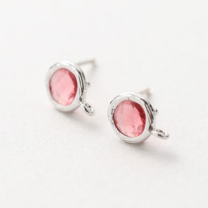 Puede incluir: Un par de pendientes de botón con una gema rosa facetada engastada en metal plateado. Los pendientes tienen un diseño orgánico con un pequeño detalle de bucle. El fondo es blanco.