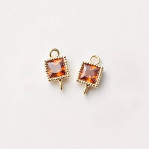 May include: Two gold-toned square-shaped charms with a small loop at the top. Each charm features a clear orange gemstone in the center.