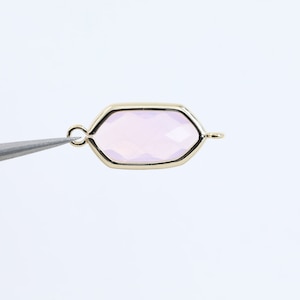 Puede incluir: Un colgante dorado con un marco hexagonal y una piedra preciosa rosa pálida en el centro. El colgante tiene un bucle a cada lado para sujetarlo a una cadena o cordón.