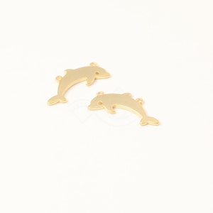 May include: Two gold-colored dolphin charms. Each charm has a curved body, a small eye, and a loop at the top for attaching to jewelry. The dolphins are facing different directions, and are set against a white background.