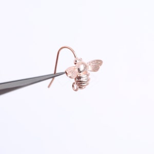 May include: A rose gold bee earring with a detailed design. The earring is held by tweezers.