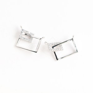 Puede incluir: Dos colgantes de plata con diseño cuadrado. Cada colgante presenta un cuadrado más pequeño dentro del cuadrado más grande, ambos incrustados con piedras de circonita cúbica transparentes.