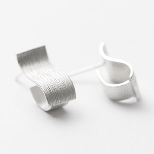 Puede incluir: Un par de pendientes de plata con un diseño ondulado texturizado. Los pendientes están hechos de un metal liso y pulido y tienen un estilo moderno y minimalista.