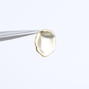 May include: A gold-coloured metal charm in the shape of a rounded, irregular circle.