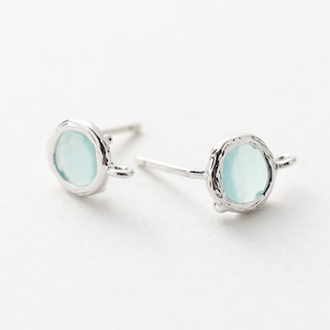 Puede incluir: Un par de pendientes de plata con una piedra redonda azul claro engastada en un marco de plata texturizado. Cada pendiente tiene un pequeño bucle de plata unido al marco.