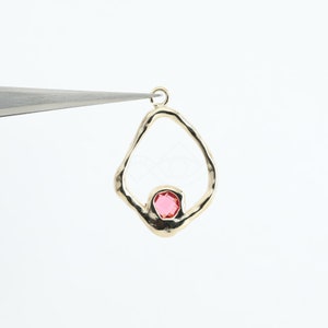 Op de afbeelding: Gouden hanger met een roze edelsteen in het midden. De hanger heeft de vorm van een traan met een onregelmatige, getextureerde rand.