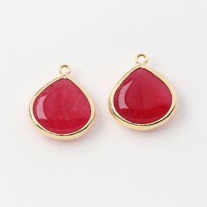 Może przedstawiać: Dwa wisiorki w kształcie łezki z czerwonymi kamieniami szlachetnymi i złotymi metalowymi obramowaniami. Każdy wisiorek ma małą pętelkę u góry do przymocowania do łańcuszka lub haczyka do kolczyków. Kamienie mają gładką, wypolerowaną powierzchnię.