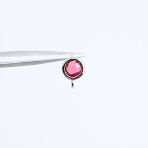 Puede incluir: Un charm plateado con un cristal redondo facetado de color rosa en el centro. El charm tiene un bucle en la parte inferior para sujetarlo a una pulsera o collar.