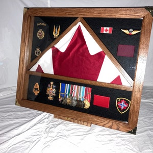 May include: A wooden display case with a folded Canadian flag, medals, patches, and insignia. The flag is red and white. The case has a black background and brass corner accents.