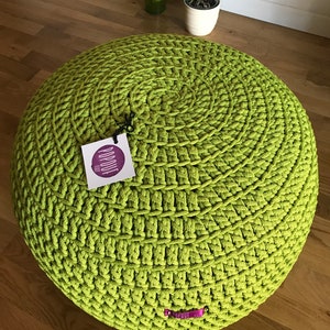Könnte beinhalten: Ein großer, runder, grüner gehäkelter Pouf-Ottomane. Der Pouf ist aus dickem, grobem Garn gefertigt und hat ein kreisförmiges Muster. Der Pouf steht auf einem Holzboden.