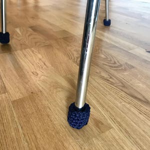 Op de afbeelding: Close-up van een stoelpot met een marineblauwe gehaakte voetbeschermer. De poot is verchroomd en reflecteert het licht. De vloer is gemaakt van houten planken.
