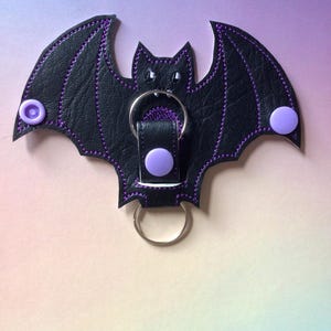 May include: Black leather bat-shaped keyring with purple stitching and accents. The bat has a metal ring and two purple button-like fasteners on the wings. The background has a gradient of purple, pink, and yellow.