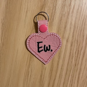May include: A pink heart-shaped keyring with the word "Ew." embroidered in black. The keyring features a button closure and a silver key ring. The edges are stitched with white and red thread.