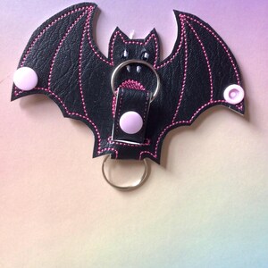 May include: A black bat-shaped keyring with pink stitching and accents. The keyring has a metal ring and two pink button closures. The bat's wings are spread, and the design is detailed with pink thread.