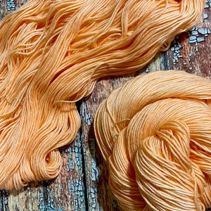 May include: A close-up of two skeins of peach-colored yarn. The yarn is soft and fluffy, and it is perfect for knitting or crocheting.
