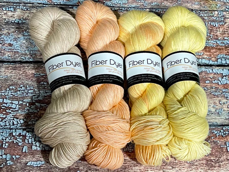 May include: Four skeins of hand-dyed yarn in shades of beige, peach, yellow, and bright yellow. The yarn is labeled "Fiber Dyed" with the company's contact information.