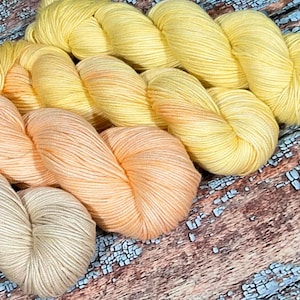 May include: Three skeins of yarn in shades of yellow, orange, and beige. The yarn is arranged in a gradient from yellow to beige.
