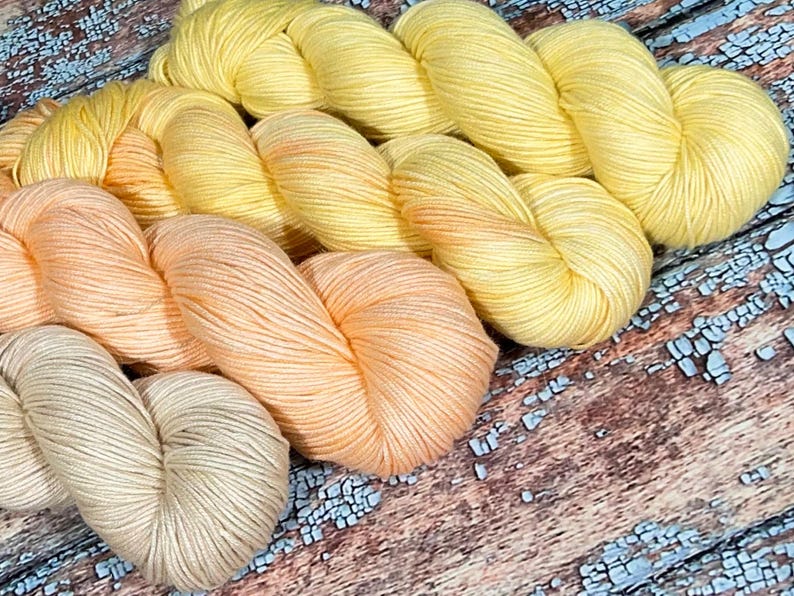 May include: Three skeins of yarn in shades of yellow, orange, and beige. The yarn is arranged in a gradient from yellow to beige.