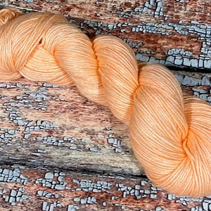 May include: A skein of peach-colored yarn, with a subtle sheen, is displayed on a rustic wooden surface.