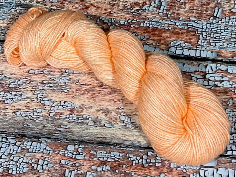 May include: A skein of peach-colored yarn, with a subtle sheen, is displayed on a rustic wooden surface.