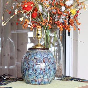 May include: A decorative table lamp with a blue and brown speckled ceramic base. The lamp has a brass-colored neck and a gold-toned lamp holder. A vase of colorful flowers sits behind the lamp.