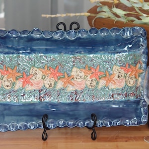 May include: A rectangular ceramic serving platter with a blue and turquoise glaze. The platter features a border of seashells and a central design of starfish and seashells. The platter is supported by a black metal stand.