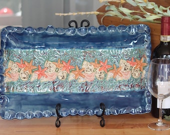 May include: A rectangular ceramic serving platter with a blue and turquoise glaze. The platter features a border of seashells and a central design of starfish and seashells. The platter is supported by a black metal stand.