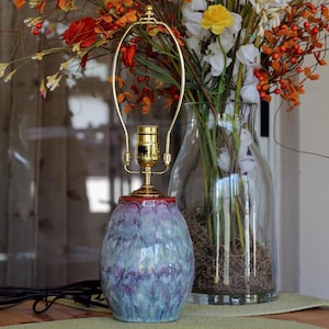May include: A table lamp base featuring a rounded ceramic body with a mottled glaze in shades of blue, purple, and red. The lamp has a polished brass socket and a harp attachment, with a black electrical cord extending from the base.