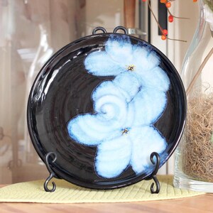 May include: A black ceramic plate with a light blue floral design, displayed on a black metal stand. The flowers have a detailed, layered appearance with yellow centers. The plate is set against a soft yellow background.