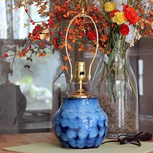 May include: A table lamp base featuring a rounded, bulbous ceramic body with a mottled blue and white glaze. The lamp has a polished gold-tone metal socket and a tall, arched wire harp extending upward from the top of the ceramic base.