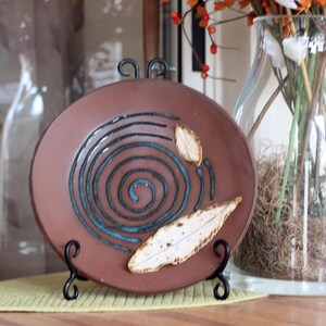 May include: A brown ceramic plate with a blue spiral design and leaf accents, displayed on a black metal stand. The plate is set on a yellow placemat, with a glass vase of flowers in the background.
