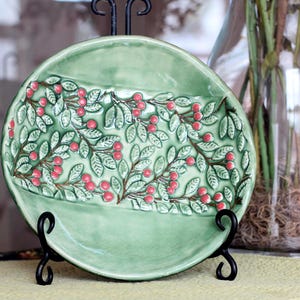 May include: A round, light green ceramic plate with a raised design of leaves and red berries. The plate is displayed on a black metal stand. The plate's design is centered and has a slight curve.